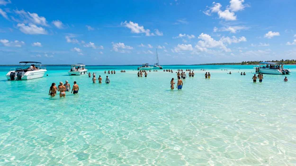 Boats anchored around a shallow sandbar in the open sea, with people standing in pale turquoise water